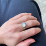 Load image into Gallery viewer, A close-up of a hand wearing a silver ring with an oval white cabochon moonstone, resting on dark navy blue fabric.
